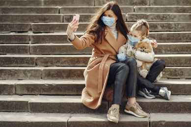 young woman hugging cute daughter with dog in masks taking selfie on steps